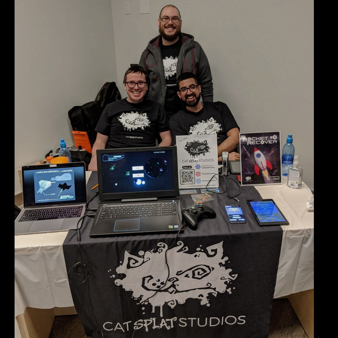 Three members of Cat Splat Studios at a convention booth in black studio shirts, with laptops running Codename: Shifter, an Xbox controller, and a Rocket Recover poster.